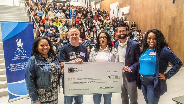 Five adults stand in front of a body of middle school students holding a promotional check. 