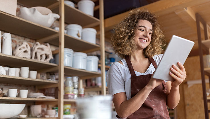 A woman in a pottery studio holds a tablet smiling. 